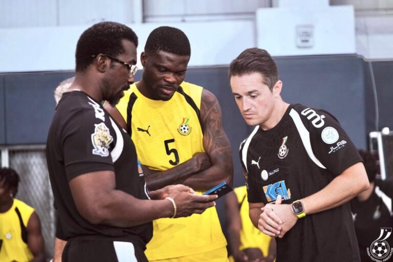 Three men in sports jerseys huddle and look at a smartphone together, likely reviewing a play or strategy.