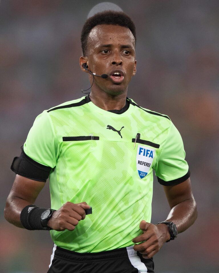 Soccer referee in a bright neon jersey with FIFA badge running on the field, wearing a headset mic and black armbands.