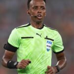 Football referee running, wearing a bright green FIFA jersey with a headset microphone and wristwatch.