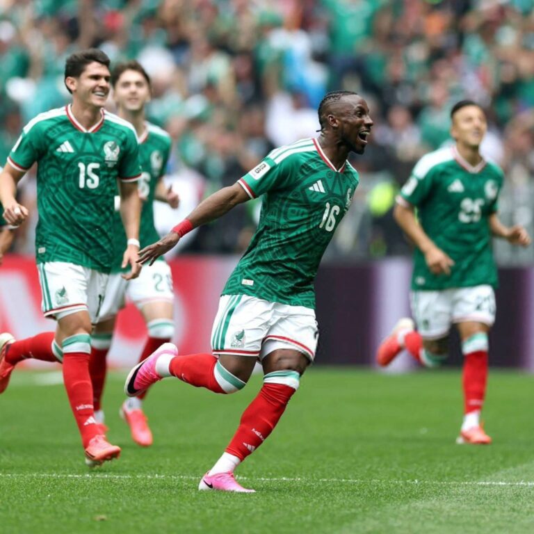 Soccer players in green jerseys sprint on a grass field during a match with a cheering crowd in the stands behind them.