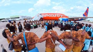 Group of people in brown patterned outfits circle the tarmac beside a bright orange Sun Country airplane, with stairs and luggage nearby.
