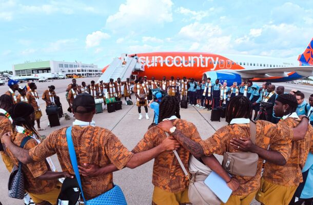 IMG_0909 | Ghanaguardian.com Group of people in brown patterned outfits circle the tarmac beside a bright orange Sun Country airplane, with stairs and luggage nearby.