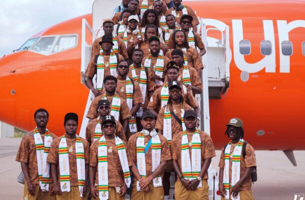 IMG_0910 | Ghanaguardian.com Group of men in brown outfits with white, green, yellow scarves pose on airplane stairs in front of an orange plane, facing camera.