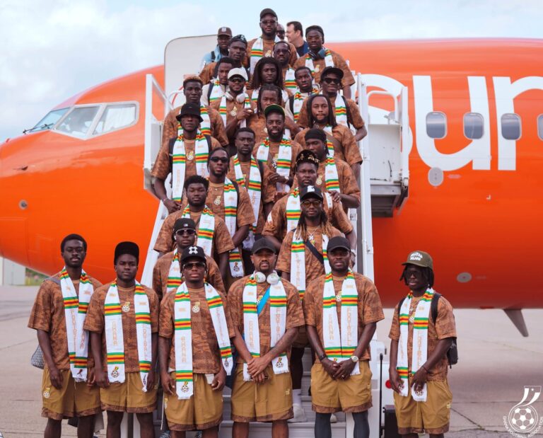 Group of men in brown outfits with white, green, yellow scarves pose on airplane stairs in front of an orange plane, facing camera.