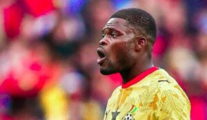 Soccer player in a yellow jersey shouting on the field with a blurred stadium crowd in the background.