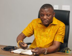 Man in a bright yellow patterned shirt sits at a desk, writing in an open notebook with a pen, wearing a bracelet and watch.