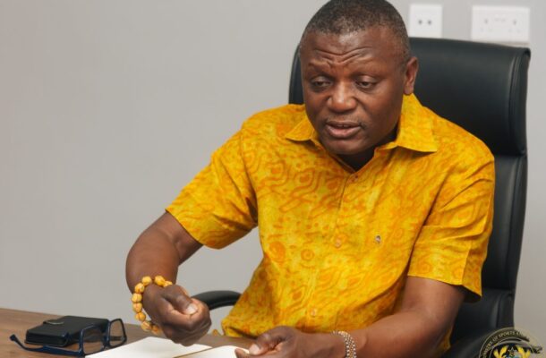 IMG_0915 | Ghanaguardian.com Man in a bright yellow patterned shirt sits at a desk, gesturing with his right hand over an open notebook.