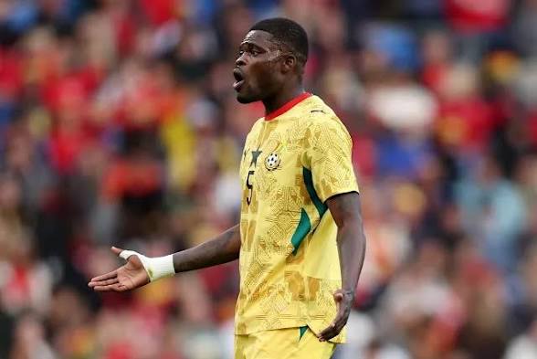 Soccer player in a yellow jersey with red collar (number 5) on the pitch, arms outstretched in a questioning gesture.