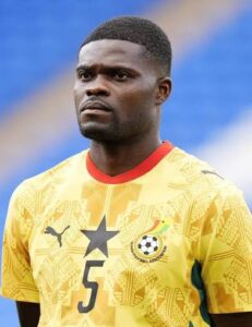 Male footballer in a yellow Ghana national team jersey with a black star emblem and number 5, standing on the field with a serious expression.