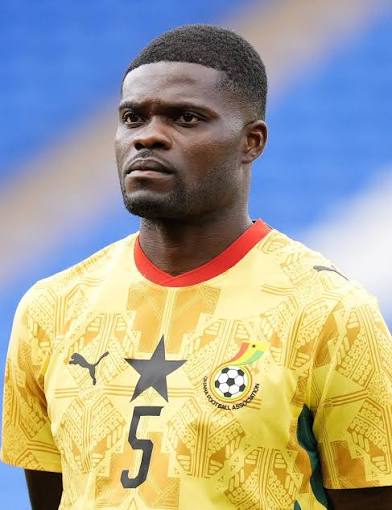 Male footballer in a yellow Ghana national team jersey with a black star emblem and number 5, standing on the field with a serious expression.