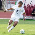 Soccer player wearing a white jersey with number 10 and a star crest, dribbling a ball on a grassy field near a bench area.