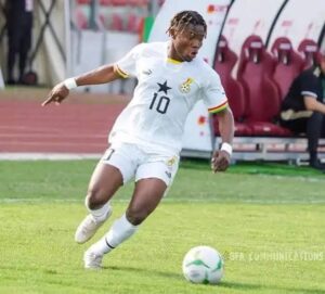 Soccer player wearing a white jersey with number 10 and a star crest, dribbling a ball on a grassy field near a bench area.