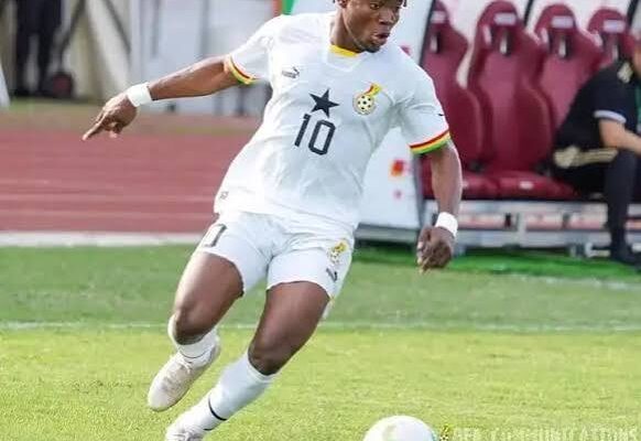 IMG_0930 | Ghanaguardian.com Soccer player wearing a white jersey with number 10 and a star crest, dribbling a ball on a grassy field near a bench area.