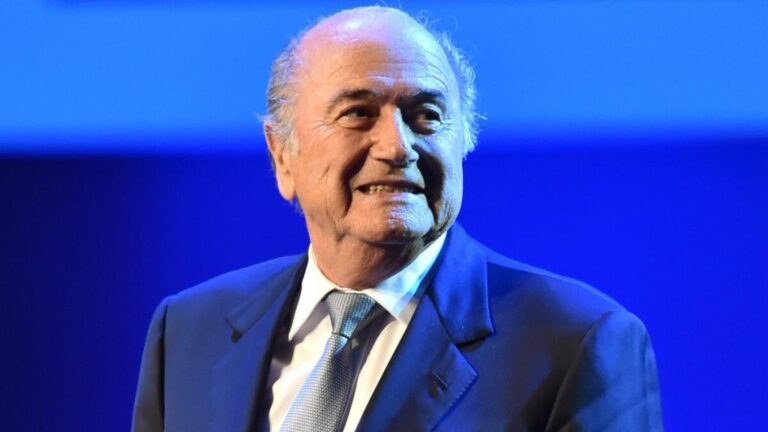 Older man in a dark suit and light blue tie smiles on a stage with a blue backdrop.