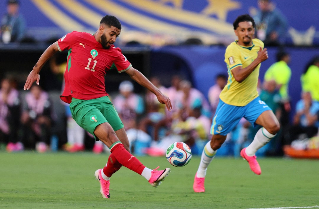 Two soccer players chase the ball on a grass field; one in red jersey number 11 with green shorts, the other in yellow jersey with blue shorts.