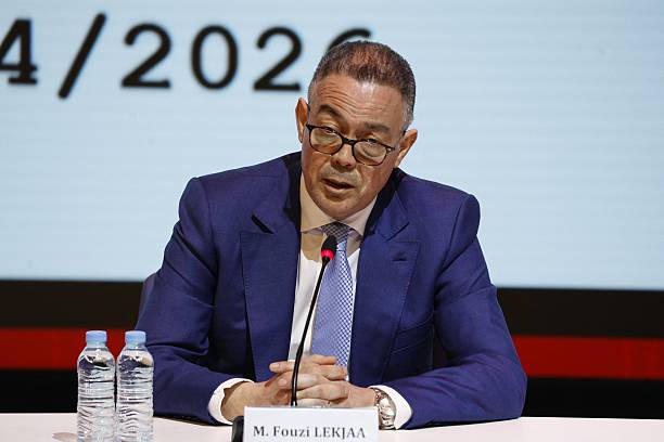 Male speaker in a blue suit sits at a panel with a microphone, flanked by two water bottles; nameplate reads 'M. Fouzi LEKJAA' (Lekjaa).