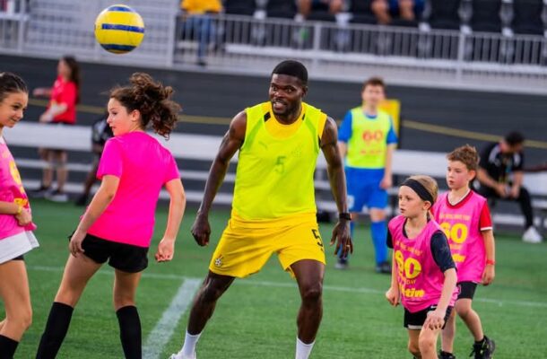 Adult soccer player in neon yellow kit controls a ball in the air while kids in pink bibs practice around him on a turf field near a stadium.