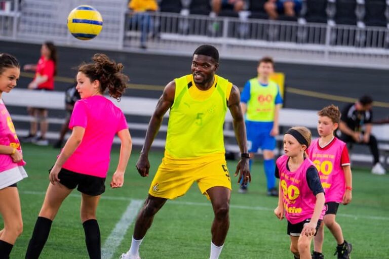 Adult soccer player in neon yellow kit controls a ball in the air while kids in pink bibs practice around him on a turf field near a stadium.