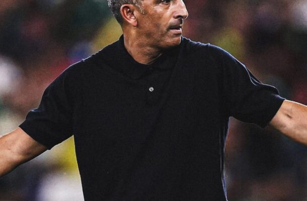 IMG_0943 | Ghanaguardian.com Football coach in a black polo extending his arms on a stadium field during a game, with blurred spectators in the background and FIFA logos on the left.
