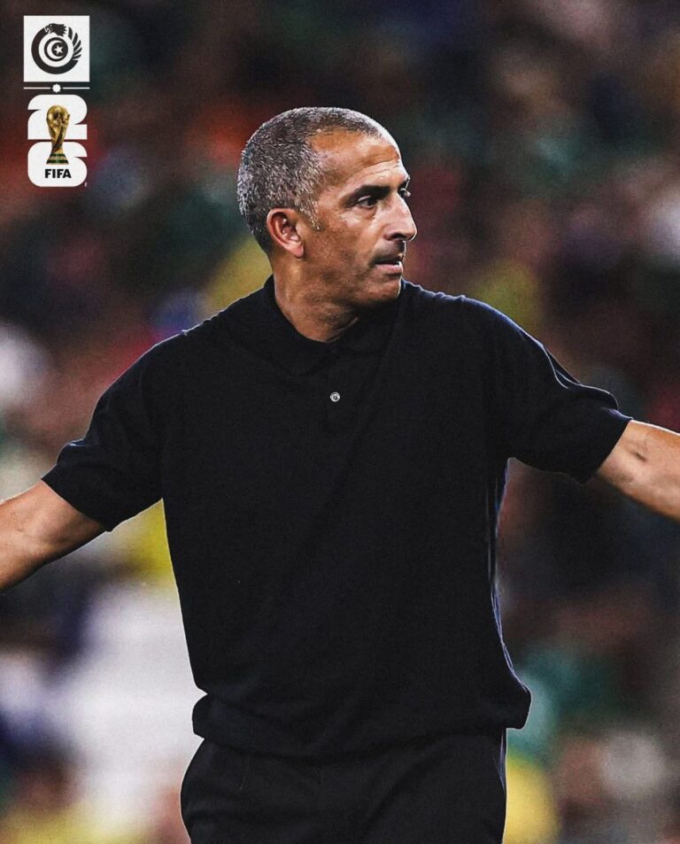 Football coach in a black polo extending his arms on a stadium field during a game, with blurred spectators in the background and FIFA logos on the left.