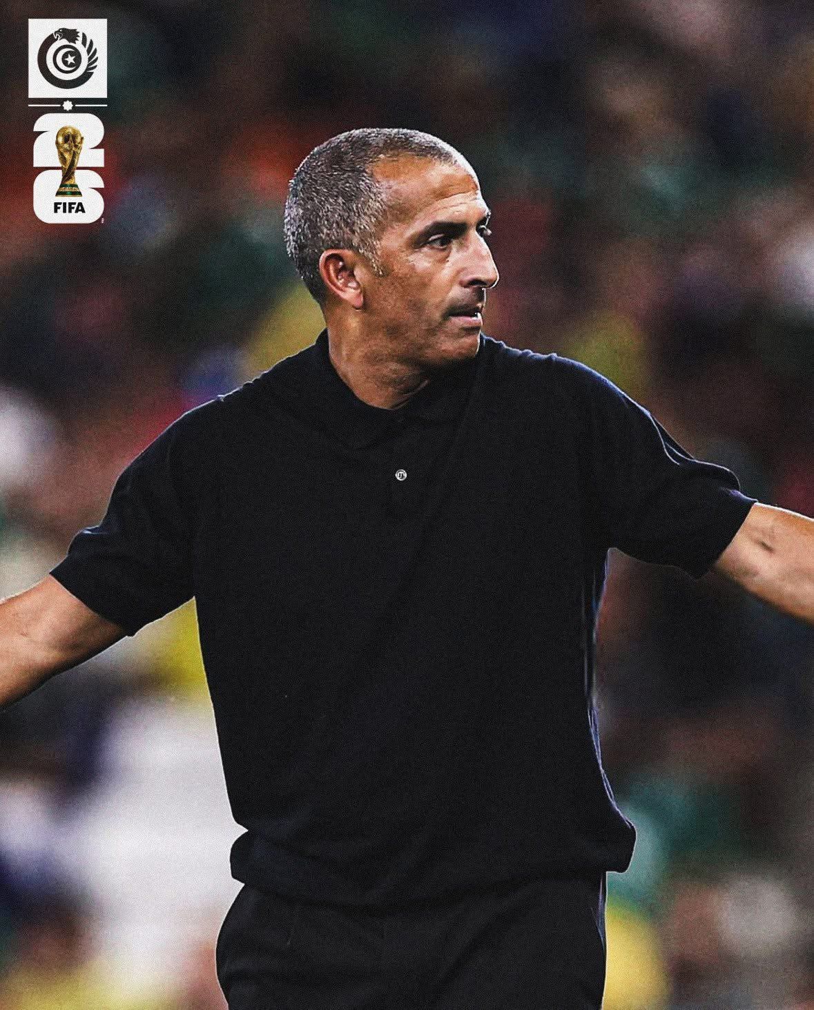 Football coach in a black polo extending his arms on a stadium field during a game, with blurred spectators in the background and FIFA logos on the left.