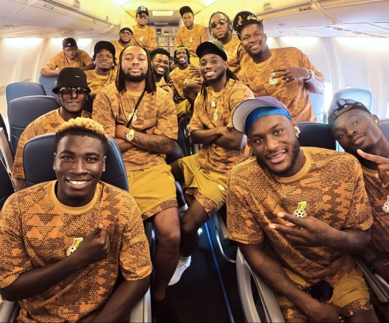 Group of men in matching brown camo shirts on a plane, posing for a team photo with smiles.