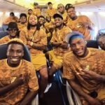 Group of athletes in matching orange camouflage shirts posing for a photo inside an airplane.