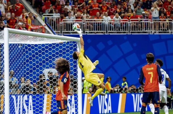 Soccer goalkeeper in yellow leaps to punch away the ball at the goal as players watch; crowded stands behind.