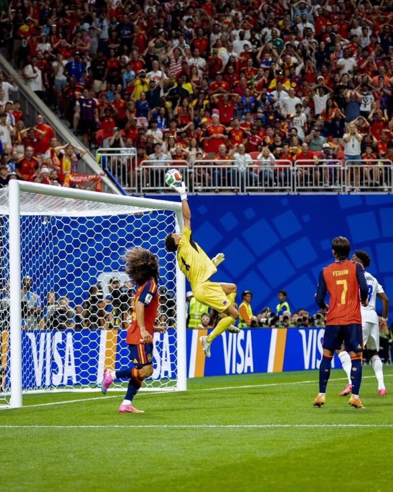 Soccer goalkeeper in yellow leaps to punch away the ball at the goal as players watch; crowded stands behind.