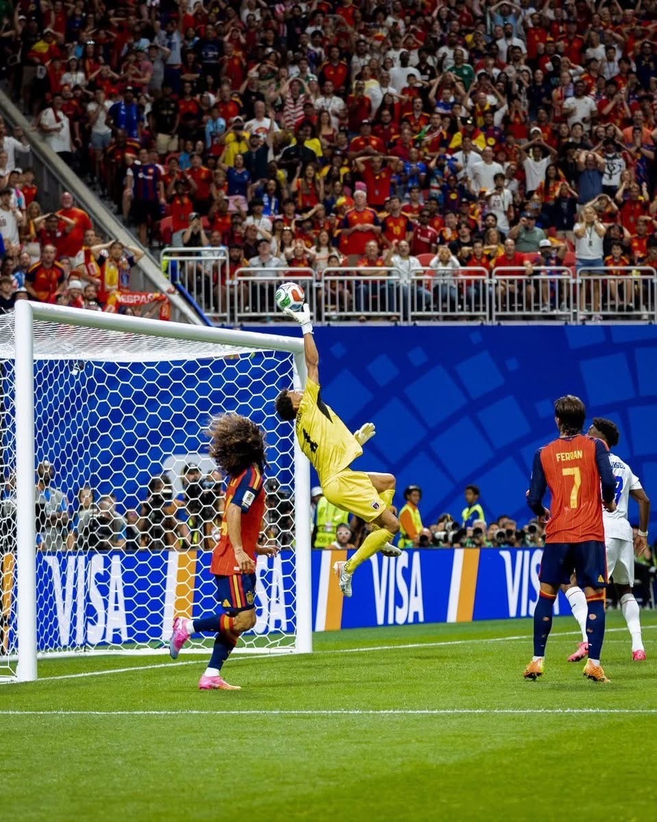 Soccer goalkeeper in yellow leaps to punch away the ball at the goal as players watch; crowded stands behind.