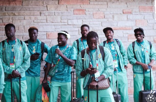 Group of young men in matching teal tracksuits with backpacks and rolling suitcases posing against a stone wall outside.