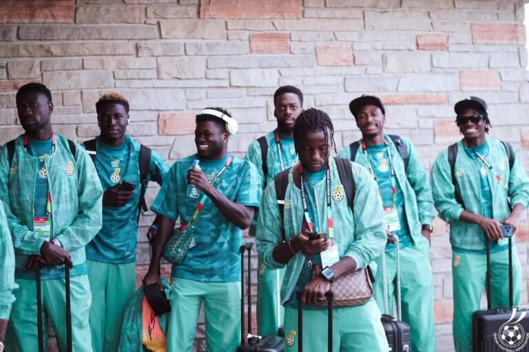 Group of young men in matching teal tracksuits with backpacks and rolling suitcases posing against a stone wall outside.