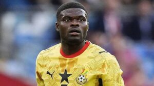 Young footballer in a yellow jersey looking surprised on the field, stadium blurred in the background with fans lined up behind him.