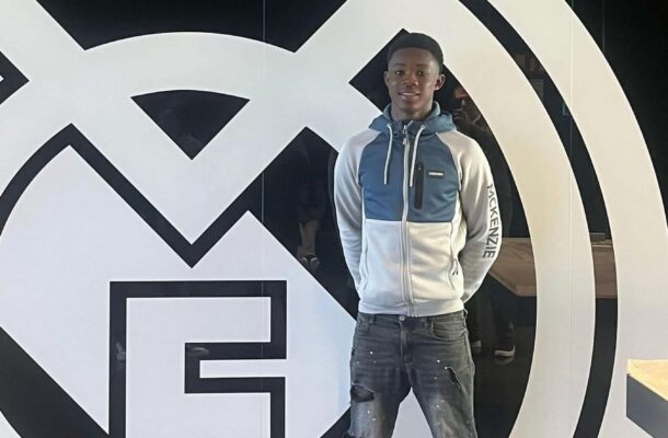 Young man in a blue-gray hoodie and ripped jeans stands in front of a large black-and-white circular logo inside a modern space.