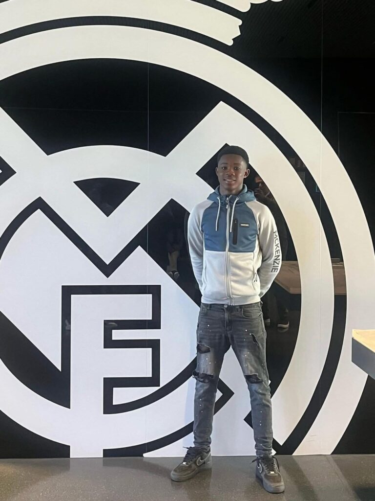 Young man in a blue-gray hoodie and ripped jeans stands in front of a large black-and-white circular logo inside a modern space.