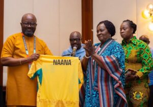 Group of four adults on stage with a yellow jersey labeled NAANA being presented or displayed.