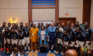 Group photo of Ghanaian football players in black jerseys with officials indoors, team posing together.