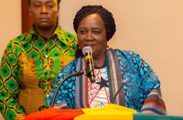 Woman in a blue patterned outfit speaks into a microphone at a podium covered with Ghana's flag colors, with a man in a green patterned shirt in the background.