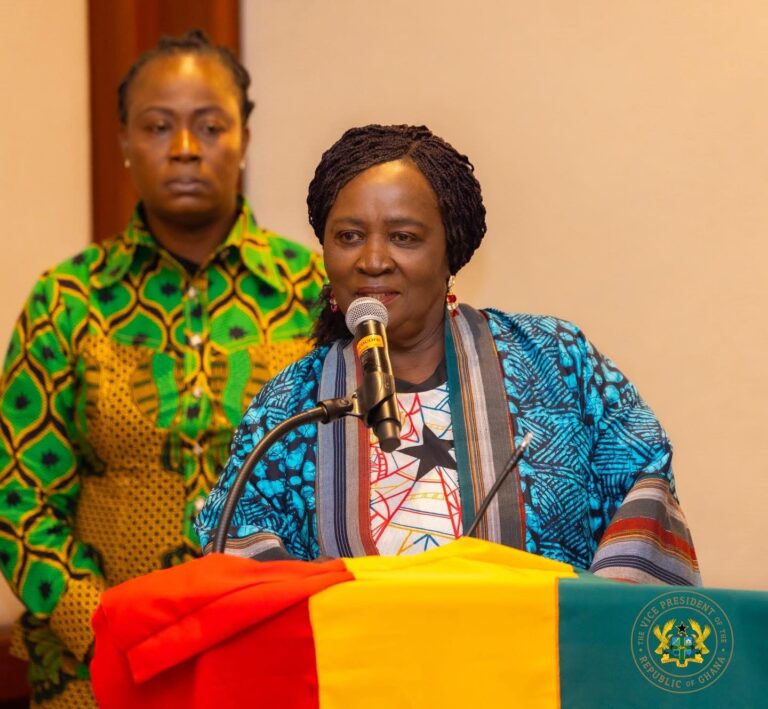 Woman in a blue patterned outfit speaks into a microphone at a podium covered with Ghana's flag colors, with a man in a green patterned shirt in the background.
