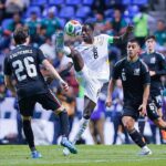 Soccer player in a white jersey with number 8 leaps to control the ball while two dark-clad defenders approach.