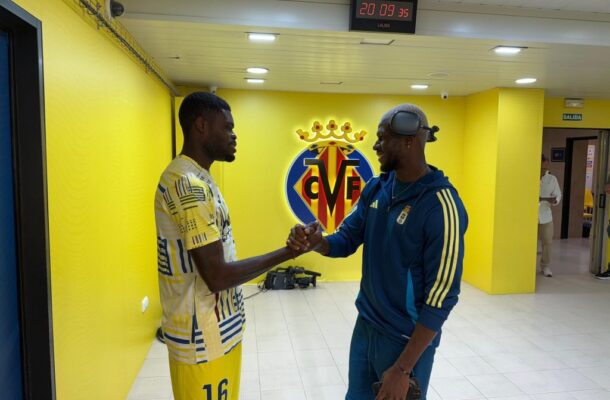 Two men shake hands in a bright yellow club corridor with a Villarreal CF crest on the wall behind them, one in a yellow kit and the other in blue tracksuit.