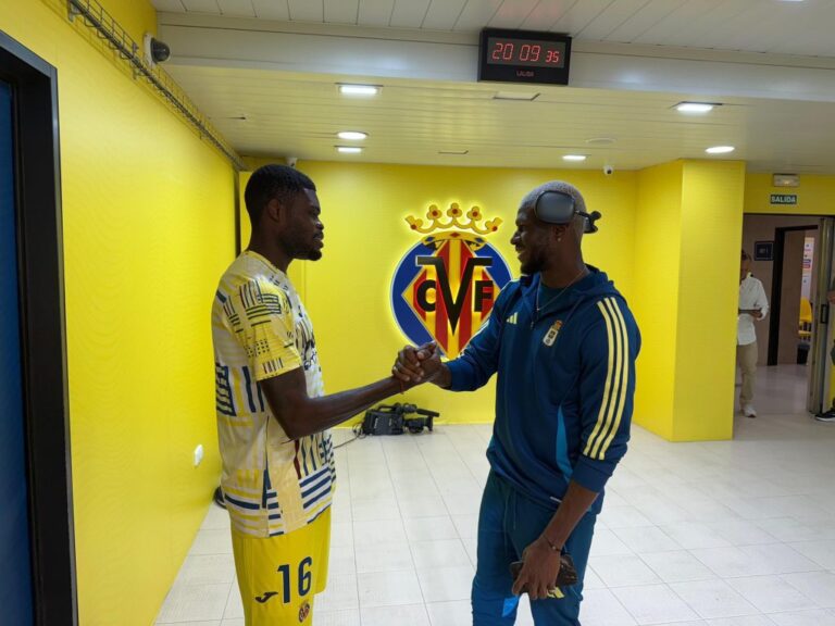 Two men shake hands in a bright yellow club corridor with a Villarreal CF crest on the wall behind them, one in a yellow kit and the other in blue tracksuit.