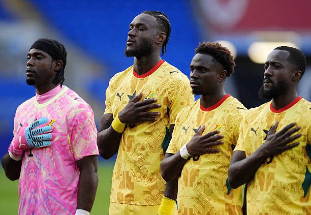 Four football players stand with hands over their hearts before a match; three wear yellow jerseys, one in a pink goalkeeper kit, on a green field.