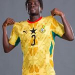 Young male soccer player posing in a yellow jersey with number 3, pulling the sides to show the shirt, against a gray backdrop.
