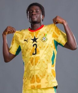 Young male soccer player posing in a yellow jersey with number 3, pulling the sides to show the shirt, against a gray backdrop.