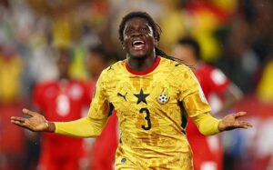 Ghana national team defender wearing yellow jersey with star and number 3, arms outstretched in celebration on a rainy stadium field