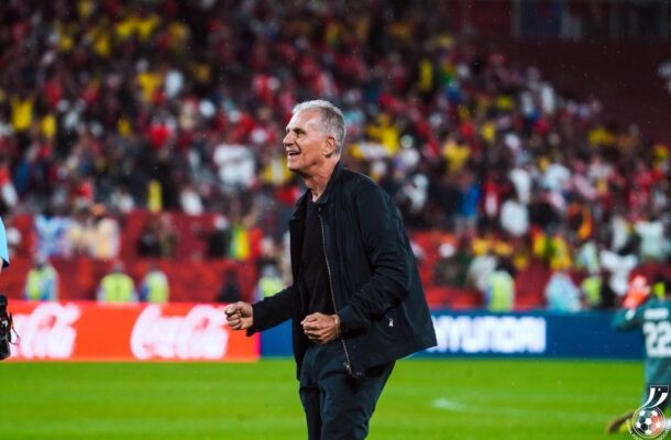 Football coach in a black jacket walking on the pitch with a cheering crowd behind him on the stands.
