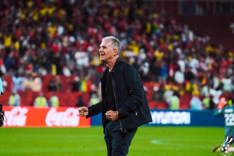 Football coach in a black jacket walking on the pitch with a cheering crowd behind him on the stands.