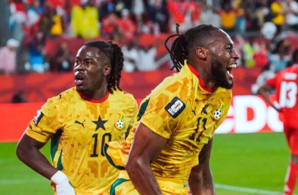 Two Ghanaian football players in yellow uniforms celebrate on the field as a cheering crowd looks on in the stands.