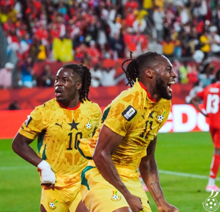 Two Ghanaian football players in yellow uniforms celebrate on the field as a cheering crowd looks on in the stands.